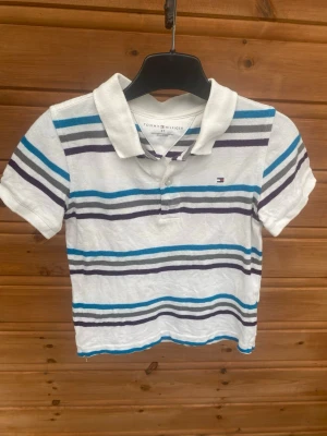 Randig pikétröja Tommy Hilfiger - Säljer en vit pikétröja från Tommy Hilfiger i storlek 4T med blå, grå och mörklila ränder. Tröjan har klassisk krage och korta ärmar, samt liten logga broderad på bröstet. Perfekt för en chill och stilren look.