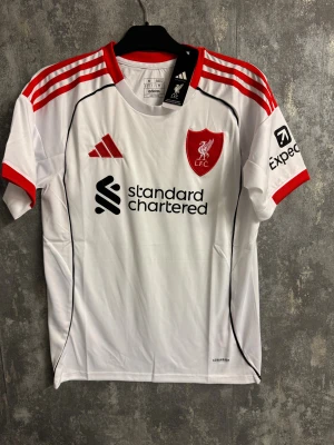 Vit Liverpool adidas matchtröja S - Säljer en officiell Liverpool FC matchtröja från adidas i storlek S. Tröjan är vit med röda detaljer, röda ränder på axlarna och klubbmärke på bröstet. Materialet är polyester och modellen är slim fit. Perfekt för dig som älskar fotboll och vill ha en snygg supportertröja.