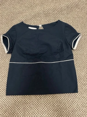 Mörkblå topp från Victoria Barrueco - Snygg mörkblå topp från Victoria Barrueco med vita detaljer längs ärmslut och midja. Kortärmad modell med rund halsringning och knapp i nacken. Perfekt för en stilren och modern look. Köpt på Sellpy.