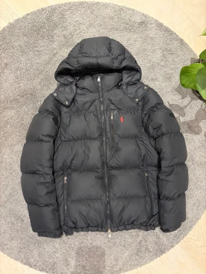 Svart dunjacka från Ralph Lauren - Snygg svart dunjacka från Ralph Lauren med huva och dragkedjor framtill. Jackan har en quiltad design och en liten röd logga på bröstet. Perfekt för kalla dagar och riktigt skön att bära. Klassisk look som funkar till det mesta.