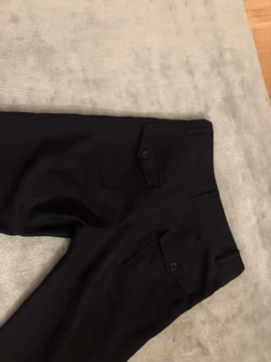 Svarta kostymbyxor från Gina Tricot - Stilrena svarta kostymbyxor från Gina Tricot med fickor bak som har lock och knapp. Byxorna har hög midja och klassisk passform, perfekta till en dressad look. Två knappar framtill och diskret dragkedja.