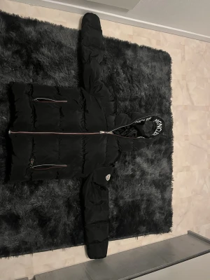 Svart Moncler dunjacka med huva - Snygg svart dunjacka från Moncler med stor huva där Moncler-loggan är tryckt runt kanten. Jackan har två dragkedjeförsedda fickor fram och Moncler-märke på ärmen. Puffig modell med quiltade partier och långa ärmar. Perfekt för kalla dagar.