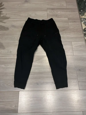 Svarta mjukisbyxor från Nike - Säljer ett par svarta mjukisbyxor från Nike med snörning i midjan och diskret Nike-logga på benet. Byxorna har muddar vid bensluten och är perfekta för chill dagar eller träning. Sköna och stilrena med normal passform.