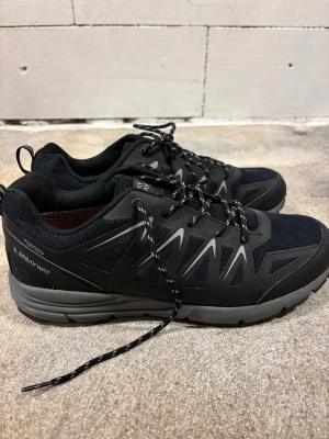 Svarta sneakers från Sprayway - Svarta sneakers från Sprayway med grå detaljer och reflekterande mönster på sidorna. Skorna har snörning, meshpaneler för andning och en robust grå sula. Insidan är orange och materialet är en mix av syntet och textil. Perfekta för träning eller vardagsaktiviteter.