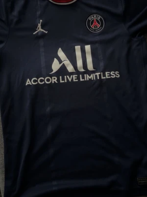 PSG x Jordan mörkblå t-shirt - Säljer en mörkblå PSG t-shirt från Jordan med vit logga och texten 'Accor Live Limitless' på bröstet. T-shirten har korta ärmar, rund hals med röd, vit och blå kant samt PSG-märke på bröstet. Perfekt för dig som gillar fotboll och streetwear.