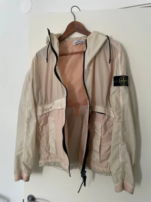 Beige vindjacka från Stone Island - Snygg beige vindjacka från Stone Island med huva och svart logotyp-patch på ärmen. Jackan har dragkedja framtill, snörning i midjan och är tillverkad i lätt syntetmaterial. Perfekt för vår och höst när du vill ha en stilren och sportig look. Går att diskutera pris vid seriöst och snabbt köp