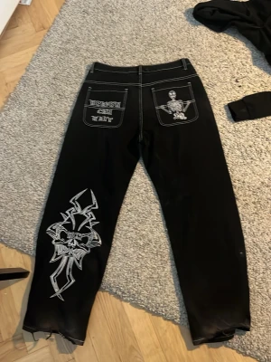 Svarta jeans med vit brodyr och tryck -  svarta heaven  jeans med vit kontrastsöm, broderad dödskalle och text på bakfickorna samt stort grafiskt tryck på ena benet baggy jeans  och normal passform. Perfekta för dig som vill sticka ut med edgy detaljer.
