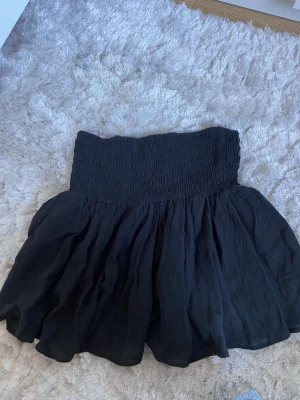 Svart kortkjol från Nelly - Säljer en svart kortkjol från Nelly med bred smock i midjan och volangig kjoldel. Kjolen har en luftig och flowy känsla, perfekt för dig som gillar en enkel men snygg stil. Passar till många olika looks!