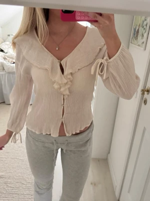 Krämvit blus med volang och knyt - Supersöt krämvit blus med volangdetaljer längs den djupa v-ringningen och knytband vid ärmsluten. Blusen har en lätt rynkad struktur och knäpps framtill. Perfekt för en romantisk och trendig look.