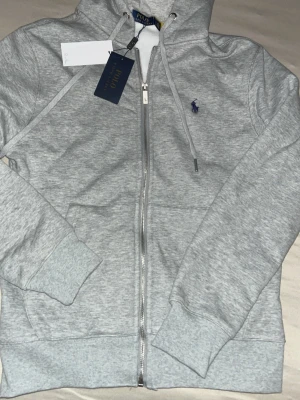 Grå hoodie från Polo Ralph Lauren - Snygg ljusgrå hoodie från Polo Ralph Lauren med dragkedja, huva och dragsnören. Klassisk blå logga broderad på bröstet. Mjuk fleece på insidan och ribbade muddar vid ärmar och nederkant. Perfekt för en chill och stilren look.