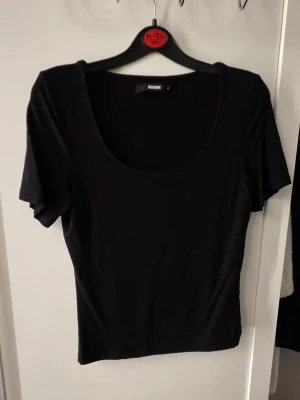 Svart topp med djup urringning Bik Bok - En enkel och stilren svart topp från Bik Bok med korta ärmar och djup, rund urringning. Tillverkad i mjukt bomullsmaterial som sitter skönt och har en normal passform. Perfekt att matcha med jeans eller kjol.