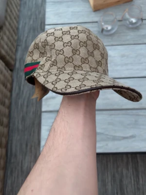 Gucci beige keps med GG-mönster - Snygg beige keps från Gucci med klassiskt GG-monogram över hela kepsen och en grön-röd rand på sidan. Tillverkad i Italien och har en böjd skärm. Materialet är canvas med bomullsfoder, perfekt för dig som gillar exklusiva accessoarer. Pris går att diskutera!