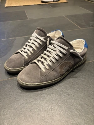 Oliver Cabell pigment 41 - Snygga grå sneakers i mocka med vit sula och vita snören. Skorna har en rund tå och en diskret detalj i form av en mörkare cirkel på sidan samt blå detalj vid hälen. Perfekta för en avslappnad och trendig stil.