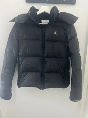 Svart pufferjacka från Calvin Klein - Snygg svart pufferjacka från Calvin Klein med huva och dragkedja framtill. Jackan har diskret CK-logga på bröstet och Calvin Klein-tryck på ärmslutet. Perfekt för kalla dagar med sin quiltade design och moderna look.