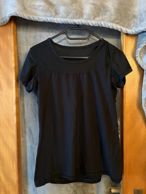 Svart topp med meshdetaljer - Snygg svart topp med korta ärmar och meshdetaljer upptill vid halsringningen och axlarna. Toppen har en enkel, rak passform och är perfekt för dig som gillar stilrena plagg med en twist.