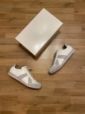 Maison Margiela White Paint Gats - Stilrena vita sneakers från Maison Margiela med gråa mockadetaljer och klassisk snörning. Skorna har en låg profil, rund tå och platt sula. Tillverkade i skinn och mocka för en lyxig känsla. Perfekta för dig som gillar minimalistisk och clean stil. Innersula 29cm, pasar då även som 45