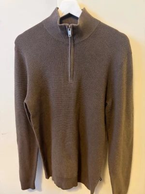 Brun stickad tröja med halv dragkedja - Säljer en brun stickad tröja med hög krage och halv dragkedja framtill. Tröjan har lång ärm och ribbade muddar vid ärmslut och nederkant. 