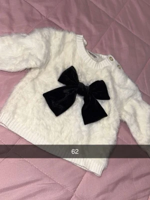 Vit fluffig stickad tröja med rosett - Supersöt vit stickad tröja med fluffig textur och stor svart sammetsrosett framtill. Tröjan har rund halsringning, ribbade muddar och knappar på axeln för enkel påklädning. Perfekt för en gullig och trendig look.