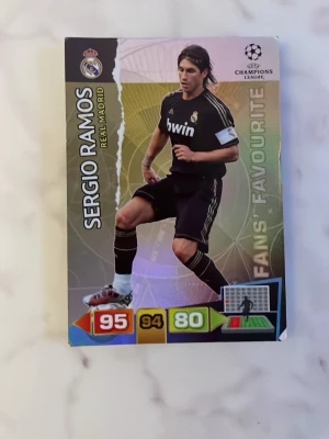 Champions League samlarkort Ramos - Samlarkort från UEFA Champions League med Sergio Ramos i Real Madrid. Kortet har färgstark design med statistik och texten 'Fans' Favourite'. Ramos bär svart matchställ och kortet har klubbmärke och turneringslogga. Perfekt för dig som gillar fotboll och samlar på sportkort.