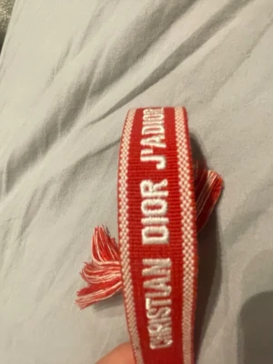 Christian Dior J'Adior rött tygarmband - Säljer ett snyggt rött tygarmband från Christian Dior med texten 'J'Adior' broderad i vitt. Armbandet har justerbar knytning med tofsar i ändarna och är perfekt för dig som gillar exklusiva accessoarer med streetstyle-vibe.