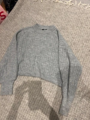 Grå croppad stickad tröja - Grå stickad tröja med croppad passform och breda ribbade ärmar. Tröjan har rund halsringning och är tillverkad i ett mjukt, melerat garn. Perfekt för lager-på-lager och enkel att styla till jeans eller kjol.