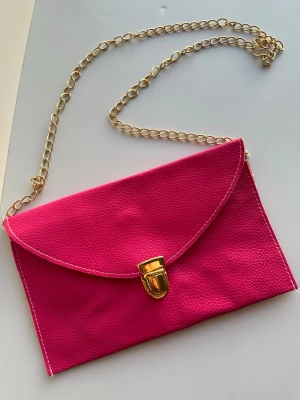 Rosa kuvertväska med guldkedja - Snygg rosa kuvertväska med guldfärgad kedja och lås. Väskan har en stilren design med struktur i materialet och vita sömmar längs kanterna. Perfekt accessoar för att lyfta din outfit.