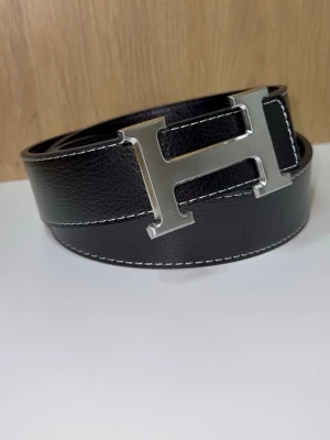 H-Bälte - Snyggt bälte med ett stort, silverfärgat H-spänne. Bältet är 115cm, har vit kontrastsöm längs kanterna och en klassisk, stilren design som lyfter vilken outfit som helst. Bältet är oanvänt (nytt). Du får även med ett armband och ett verktyg för att göra egna hål! Skriv vid minsta lilla fundering!