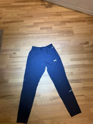 Grå mjukisbyxor från Under Armour - Snygga grå  mjukisbyxor från Under Armour med elastisk midja och logga på låret. Byxorna har en tapered passform och är perfekta för träning eller chill. Tillverkade i ett mjukt syntetmaterial som känns skönt mot huden.
