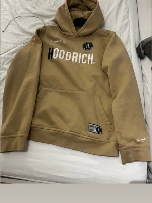 Beige hoodie från Hoodrich - Snygg beige hoodie från Hoodrich med stor logga framtill och broderad text på ärmarna. Har en klassisk huva, magficka och tryck även på ryggen. Perfekt för en streetwear-look. Mjuk bomullsblandning som känns skön mot huden.