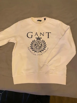 Vit sweatshirt från Gant - Säljer en stilren vit sweatshirt från Gant med stor logga och emblem på bröstet. Tröjan har rund halsringning, ribbade muddar vid ärmslut och nederkant samt en klassisk relaxed fit. Perfekt för en casual och sportig look. Bara att skieva vid frågor och priset går såklart att diskutera!