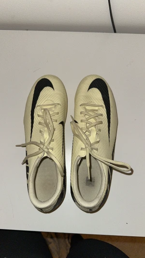 Beigea Nike fotbollsskor med snörning - Snygga beigea fotbollsskor från Nike med svart logga och snörning. Skorna har en låg profil och är designade för att ge bra grepp på planen. Tillverkade i syntetmaterial för lätt känsla och smidig passform.