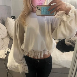 Vit blus med spetsdetaljer - Supersöt vit blus med vida ärmar och vackra spetsdetaljer vid ärmslut och nederkant. Blusen har en croppad passform och romantisk känsla, perfekt att styla med jeans eller kjol. Materialet känns mjukt och luftigt.