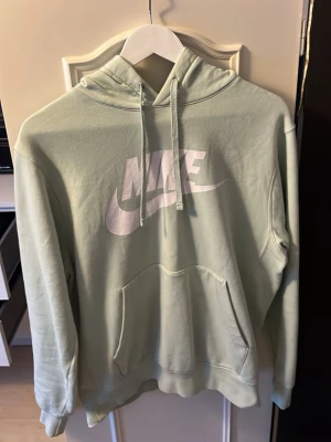 Ljusgrön Nike hoodie med logga - Snygg ljusgrön hoodie från Nike med stor vit logga på bröstet. Klassisk känguruficka framtill, justerbar huva med dragsko och mjukt material som känns skönt mot huden. Perfekt för en avslappnad och sportig stil.
