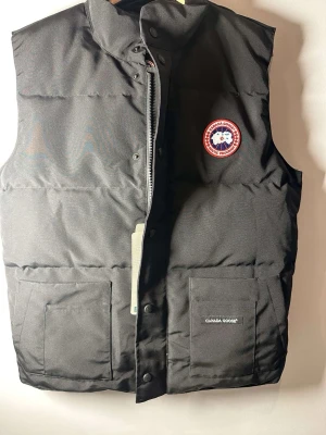 Svart dunväst från Canada Goose - Snygg svart dunväst från Canada Goose med klassisk logotyp på bröstet och två stora fickor framtill. Västen har hög krage och dragkedja med tryckknappar. Perfekt för lager-på-lager och håller dig varm under kyliga dagar.