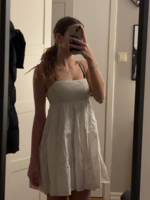 Vit kort sommarklänning - Söt vit kortklänning med smock upptill och tunna axelband. Klänningen har en luftig passform och volangig kjol, perfekt för varma dagar. Enkel och stilren design som passar till många tillfällen.