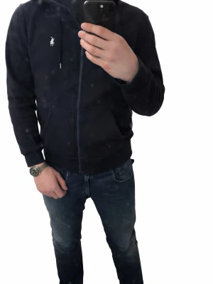 Mörkblå hoodie från Polo Ralph Lauren - Mörkblå hoodie med dragkedja från Polo Ralph Lauren. Klassisk design med huva, dragsnören och två fickor framtill. Ikoniska loggan broderad på bröstet. Perfekt för en avslappnad och stilren look.