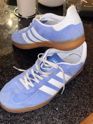 Adidas Gazelle blå/vita sneakers - Säljer ett par Adidas Gazelle sneakers i ljusblå mocka med vita detaljer och klassiska tre ränder på sidan. Skorna har vit hälkappa, vita snören och en brun gummisula. Perfekta för dig som gillar retrostil och vill sticka ut med färg.