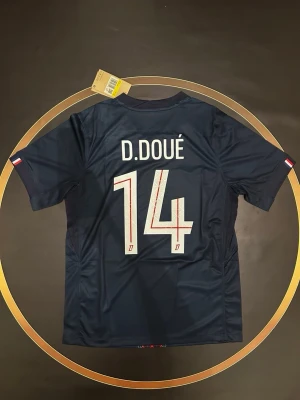 Mörkblå fotbollströja D. Doué 14 - Mörkblå fotbollströja med D. Doué och nummer 14 på ryggen. Tröjan har korta ärmar och små franska flaggor på ärmsluten. Perfekt för dig som älskar fotboll och vill bära din favoritspelares namn. S och L