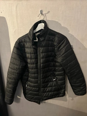 Svart dunjacka med dragkedja - Stilren svart dunjacka med quiltad design och hög krage. Jackan har dragkedja framtill och två sidofickor med dragkedja. Perfekt för kyliga dagar och enkel att matcha med olika outfits.