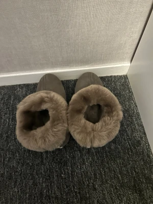 Beiga tofflor med fuskpäls - Mysiga tofflor i beige färg med mjuk fuskpäls runt öppningen. Utsidan ser ut att vara i mockaimitation och insidan är fodrad för extra värme. Perfekta för kalla dagar hemma.