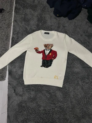 Vit stickad tröja med björn från Ralph Lauren - Vit stickad tröja från Ralph Lauren med ikonisk björn i smoking och röd kavaj på bröstet. Rund halsringning och ribbstickade muddar vid ärmslut och nederkant. RL-broderi i gult nedtill. Perfekt för dig som gillar klassiska och lekfulla plagg.