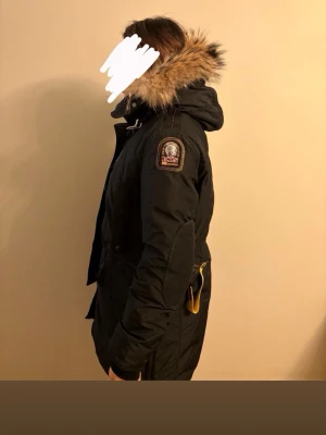 Svart Parajumpers parkas med pälskrage - Svart parkajacka från Parajumpers med ikonisk patch på ärmen, stor huva med fluffig pälskant och gul detalj vid halsen. Jackan har flera fickor, dragkedja och knappar framtill. Perfekt för kalla dagar med sin varma och rejäla design. Sparsamt använd, storlek L men passar även M. Säljer pga den ej används flera vintrar och har ingen användning av den.