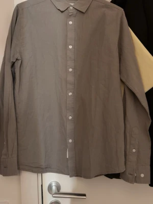 Grå skjorta med knappar - En stilren grå skjorta med klassisk krage och vita knappar framtill. Skjortan har lång ärm och är tillverkad i ett lätt bomullstyg, perfekt för en clean och enkel look.