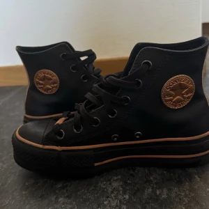 Svarta Converse Chuck Taylor High - Snygga svarta Converse Chuck Taylor high sneakers med bruna detaljer och klassisk rund logotyp på sidan. Skorna har snörning och platt sula, tillverkade i canvas med gummidetaljer. Perfekta för en cool och avslappnad stil.