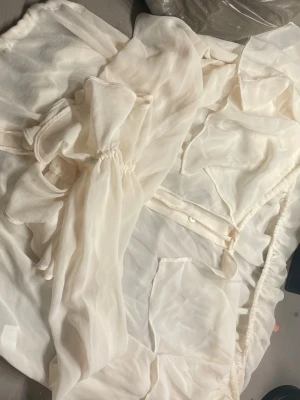 Vit blus med rosett från Topshop - Säljer en superfin vit blus från Topshop i storlek 38. Blusen har en stor rosett framtill, knappar och är gjord i ett lätt, transparent material som känns luftigt och elegant. Perfekt att styla med jeans eller kjol för en chic look.