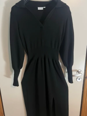 Svart långärmad klänning från Vila - Stilren svart klänning från Vila med lång ärm, markerad midja och v-ringad hals. Klänningen har en slits framtill och är i ett mjukt stickat material som ger en snygg passform. Perfekt för dig som gillar klassisk och enkel stil.