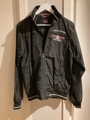 Svart vindjacka från Vinson Original - Snygg svart vindjacka från Vinson Original med broderad logga och text på bröstet. Jackan har dragkedja framtill, ribbade muddar med vita detaljer och en klassisk krage. Perfekt för dig som gillar sportig och stilren look.