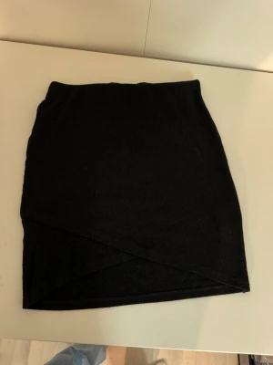 Mörkgrå omlott kortkjol i ribbad trikå - En mörkgrå kortkjol /pennkjol med omlott-design och ribbad struktur. Kjolen har en enkel och stilren look som passar perfekt till många olika outfits. Tillverkad i mjuk trikå för en bekväm känsla.🤎