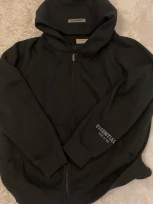 Svart hoodie från Essentials - ( Helt ny ) Svart zip-hoodie från Essentials Fear of God med tryck på ärmen. Tröjan har huva, dragkedja framtill och ribbade muddar. Perfekt för en avslappnad streetwear-look och tillverkad i mjukt material för extra komfort.