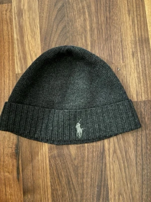 Svart stickad mössa från Polo Ralph Lauren - Snygg svart stickad mössa från Polo Ralph Lauren med klassisk ribbad kant och broderad logga framtill. Perfekt för att hålla värmen och ge en clean look. Materialet känns mjukt och följsamt, och mössan har en enkel, tidlös design.